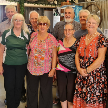 Group of nine smiling adults standing indoors, dressed in casual clothes, with rustic corrugated walls and greenery in the background.